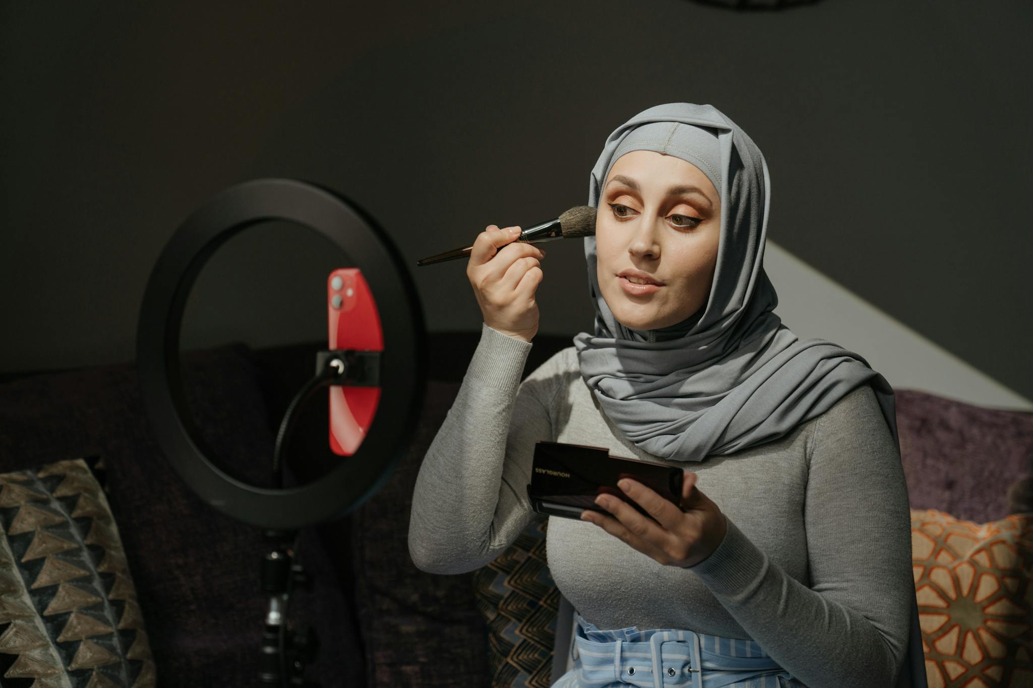 Our Mission 1 A woman in a hijab applies makeup in a beauty tutorial setting with a ring light.